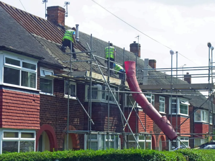 Roofers in hi-vis stripping and re-roofing a row of semi-detached houses, with a rubble chute from the scaffold
