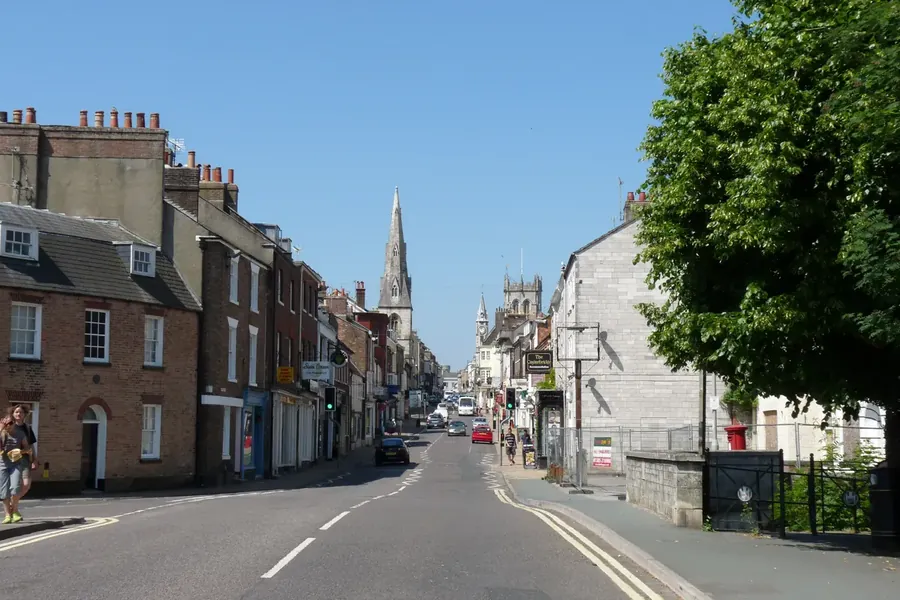 Terraced shops and houses with pitched roofs and chimneys on Dorchester's high street, with church towers beyond